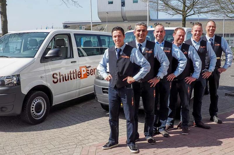 Sechs uniformierte Fahrer in Weste stehen vor zwei weißen Shuttlefahrzeugen mit Schriftzug.