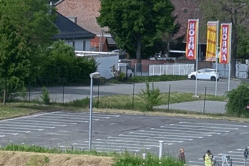 Gezäunter, größtenteils leerer Parkplatz mit markierten Stellflächen, Straßenlaterne, Werbefahnen und zwei Personen am Rand.