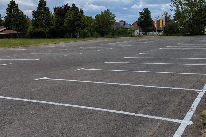 Leerer Asphaltparkplatz mit weißen Markierungen und Bäumen am Rand.