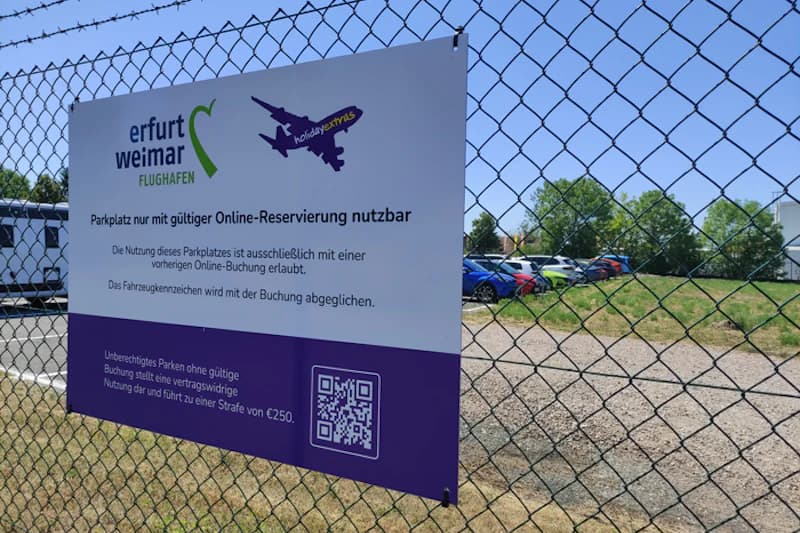 Metallzaun mit Schild des Flughafens Erfurt-Weimar und QR-Code, dahinter geparkte Autos auf eingezäuntem Gelände.
