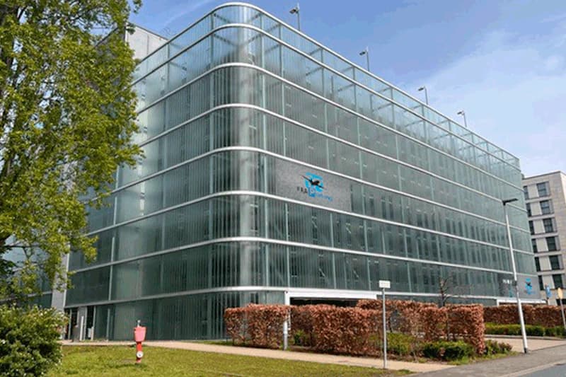 Mehrstöckiges Parkhaus mit transluzenter Glasfassade, abgerundeter Ecke, Hecke und Baum im Vordergrund.