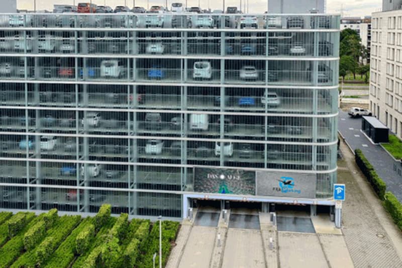 Mehrstöckiges Parkhaus mit sichtbaren Autos hinter Gitterfassade und drei Einfahrtsrampen, vor begrünten Hecken.