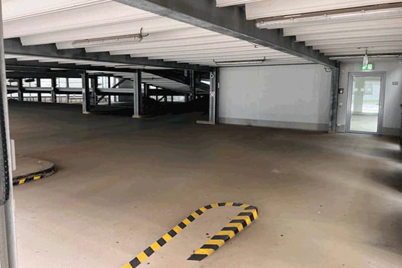 Leeres Parkhausinnenraum mit niedriger Decke, Rampe und gelb-schwarzen Schutzbaken sowie einer Notausgangstür.