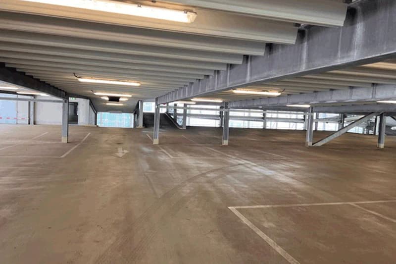 Leeres mehrgeschossiges Parkdeck mit Betonboden, Markierungen, Stahlträgern, Deckenleuchten und Rampe im Hintergrund.