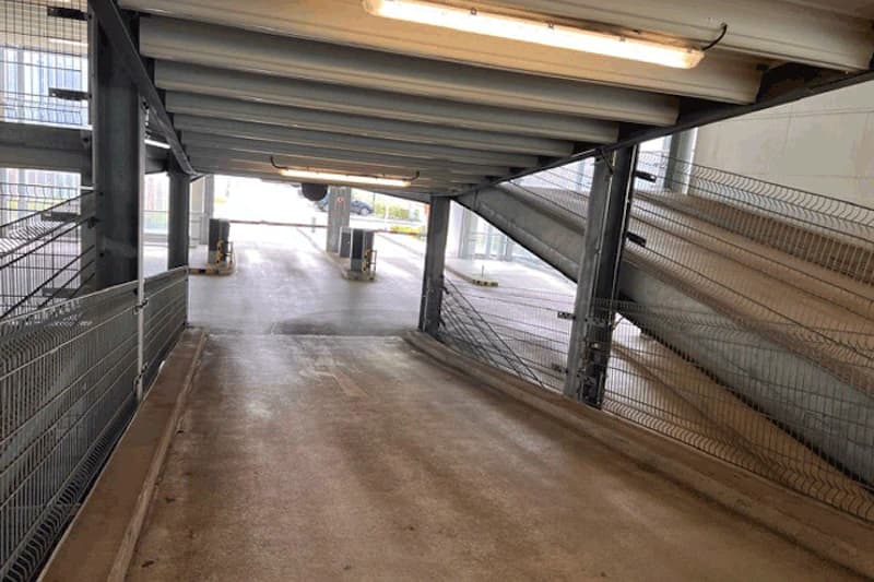 Innere Fahrspur eines Parkhauses mit abfallender Rampe, Metallgeländern, Deckenleuchten und Schranken am Ausgang.