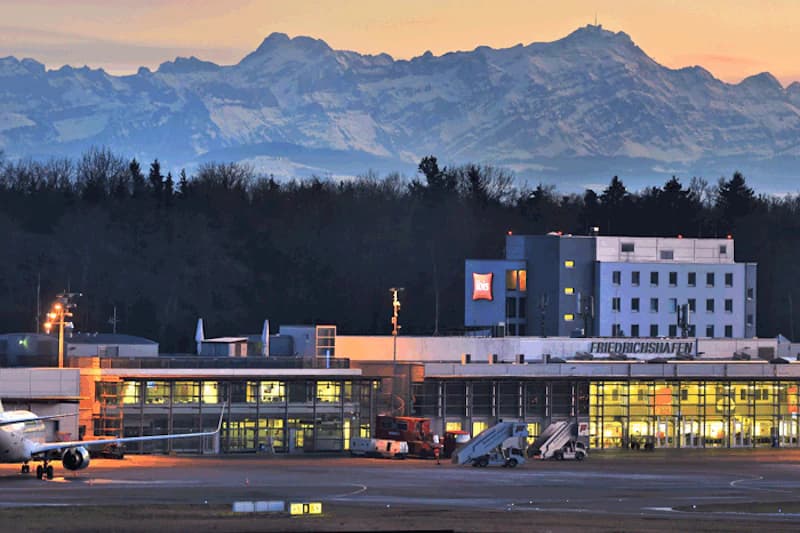 Flughafengebäude Friedrichshafen mit Flugzeugbug, Passagiertreppen und Alpen im Abendlicht.