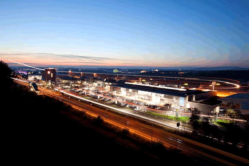 Abendliche Panoramaansicht des Flughafengeländes in Friedrichshafen mit beleuchtetem Terminal, Parkplätzen und Startbahn.