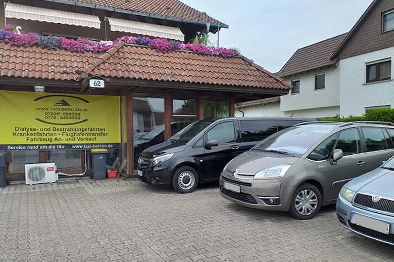 Vorgelagerter Parkplatz vor Gebäude mit gelbem Taxi Bercan Banner, drei geparkte Fahrzeuge und Blumen auf dem Dachrand.