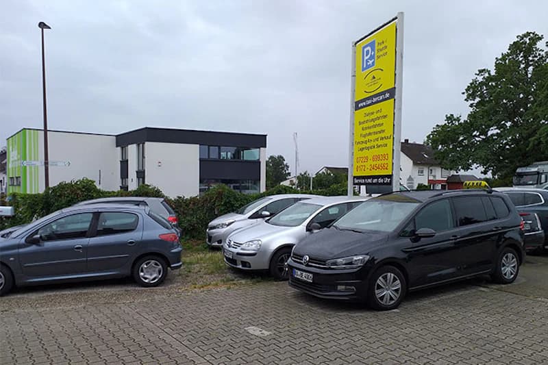 Parkplatz mit mehreren geparkten Autos, großem gelben Hinweisschild und Bürogebäude im Hintergrund.