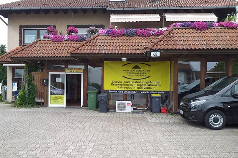 Eingangsbereich mit überdachtem Vorbau, gelbem Taxi-Bercan-Banner, Blumen am Dachrand und schwarzem Van auf gepflastertem Hof.