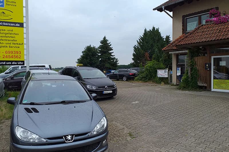 Parkplatz mit mehreren geparkten Autos, Taxi mit Dachzeichen, gelbe Werbetafel mit taxi-bercan.de und Haus mit Ziegeldach.