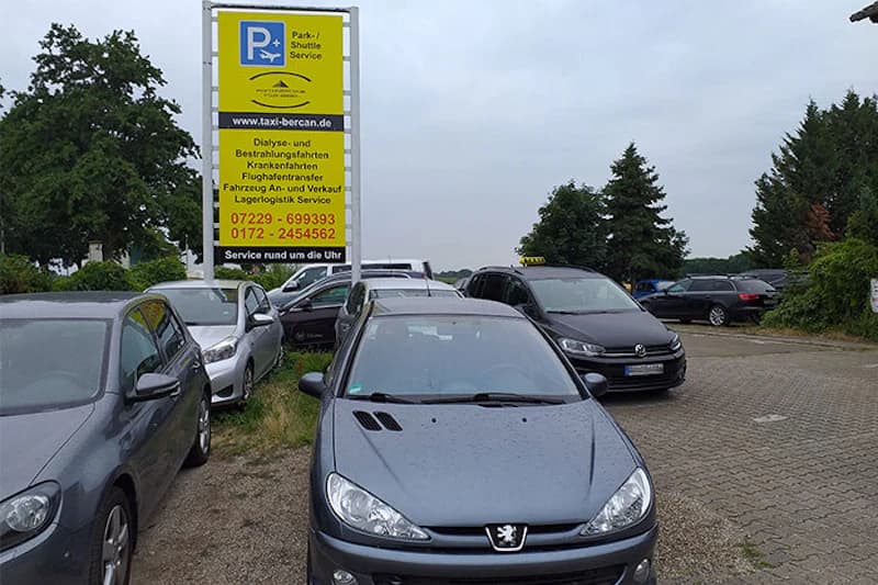 Mehrere geparkte Autos vor einem hohen gelben Schild für Park- und Abholservice am Flughafen Baden-Baden.