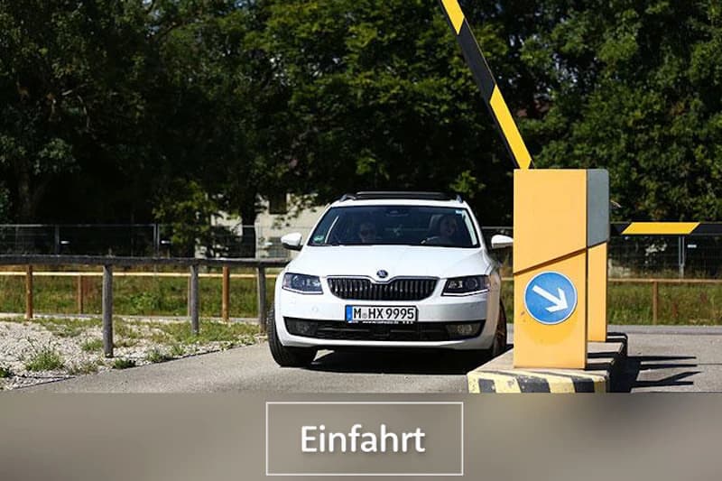 Weißes Auto hält an Schranke neben gelbem Einfahrtsautomaten mit blauem Pfeilschild.