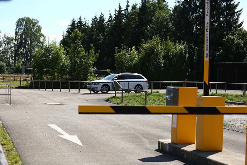 Schranke und gelbe Automaten am Zufahrtsbereich eines Parkplatzes, im Hintergrund ein weißer Kombi und Bäume.