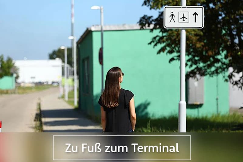 Frau von hinten auf Fußweg vor grünem Gebäude und Wegweiser mit Fuß- und Flugzeugsymbol.