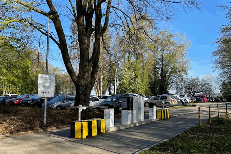 Parkplatz mit Einfahrtsschranke und Parkautomaten, geparkte Fahrzeuge und Bäume im Hintergrund.