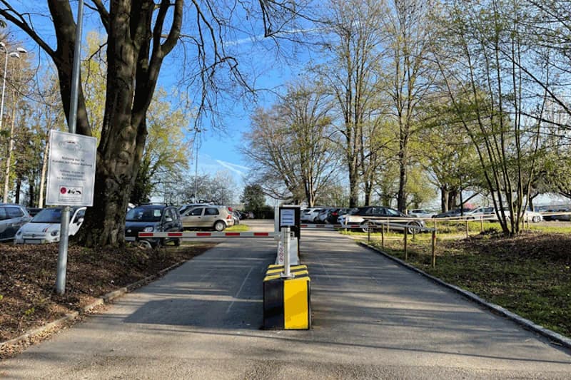 Einfahrt zu einem Parkplatz mit Schranke, Ticketautomat in Gelb, parkenden Autos und Bäumen im Hintergrund.