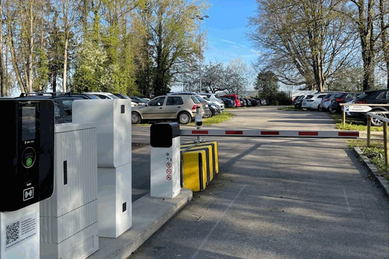 Einfahrt mit Schranke und Parkautomaten links, dahinter geparkte Autos und Baumreihe.