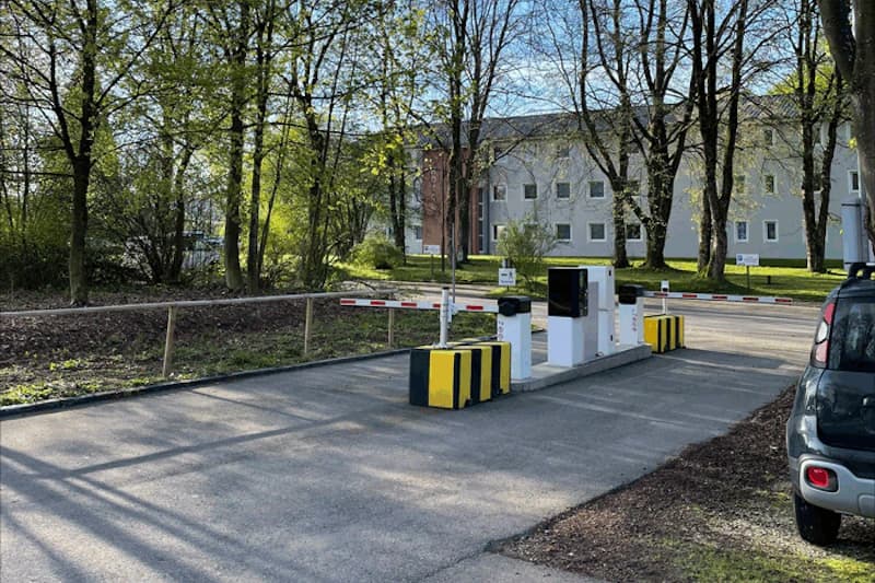Parkplatzzufahrt mit Schranke, Ticketautomaten und gelb-schwarzen Pollern vor Bäumen und Gebäude.