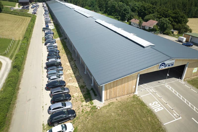 Luftbild einer langen Parkhalle mit grauem Dach, links eine lange Reihe geparkter Autos am Straßenrand.