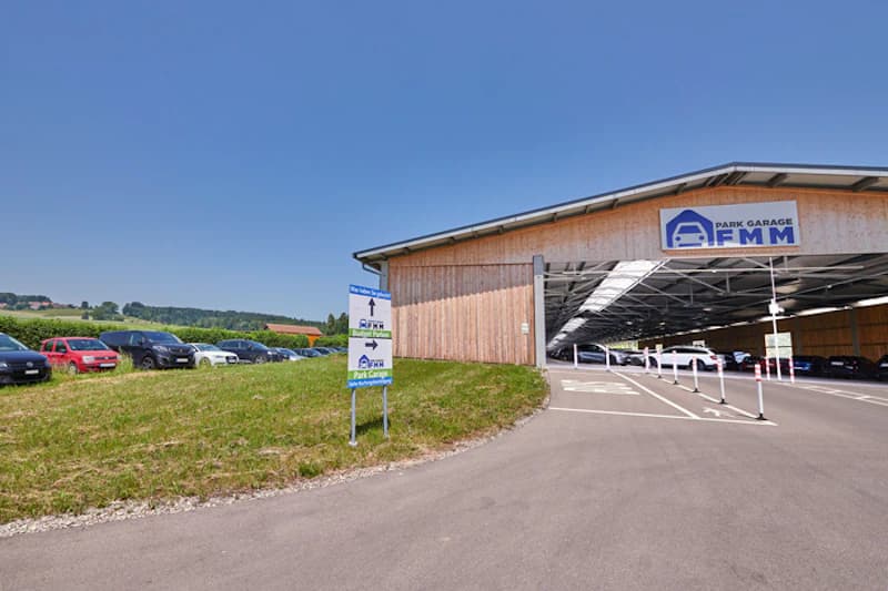 Wegweiser an einer Zufahrt vor einer offenen Parkhalle mit Holzfassade und Schild FMM.