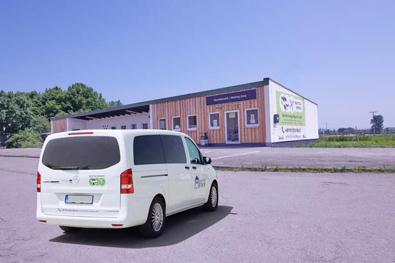 Weißer Kleinbus mit Shuttle-Aufschrift vor einem flachen Gebäude mit Holzfassade.