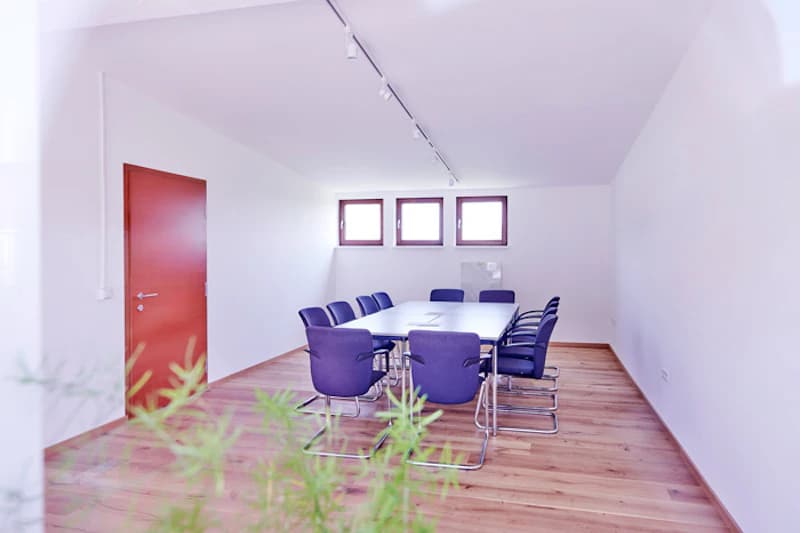 Heller Raum mit langem Tisch, blauen Stühlen, roter Tür und Holzboden.