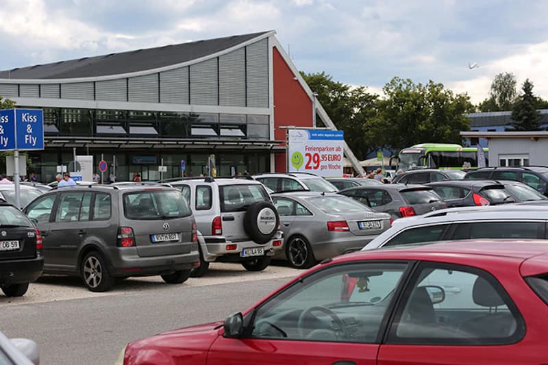 Viele geparkte Autos vor dem Terminalgebäude mit rotem Giebel und großem Werbebanner.