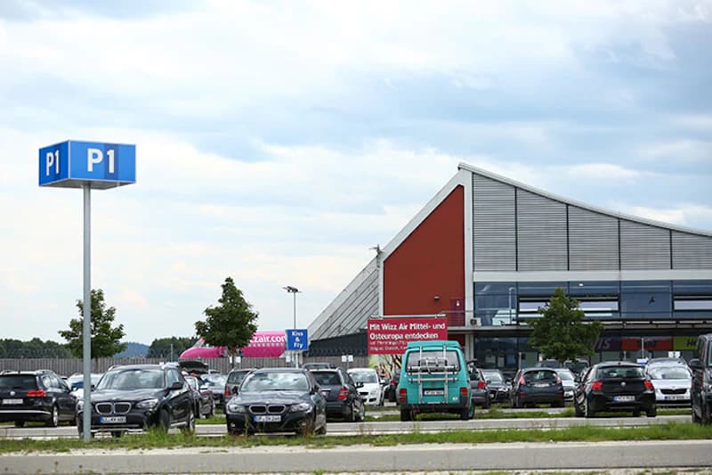 Blaues Schild P1 auf einem Mast über geparkten Autos, dahinter das Terminal mit rotem Giebel.