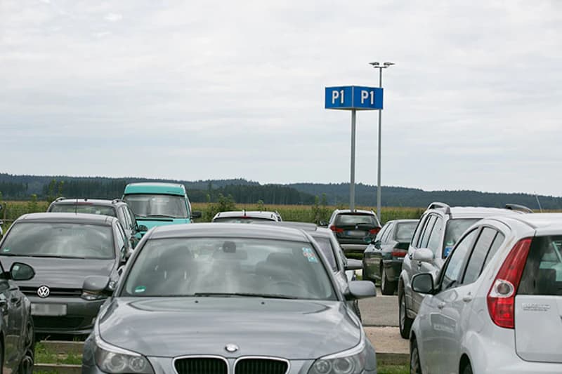Geparkte Autos auf einem Außenparkplatz, dahinter ein blaues Schild P1 auf hohem Mast und Felder.