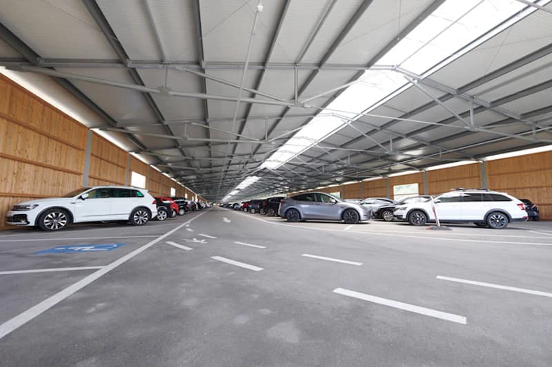 Innenraum einer hohen Parkhalle mit Stahldachträgern, Holzwänden und geparkten Autos in Reihen.