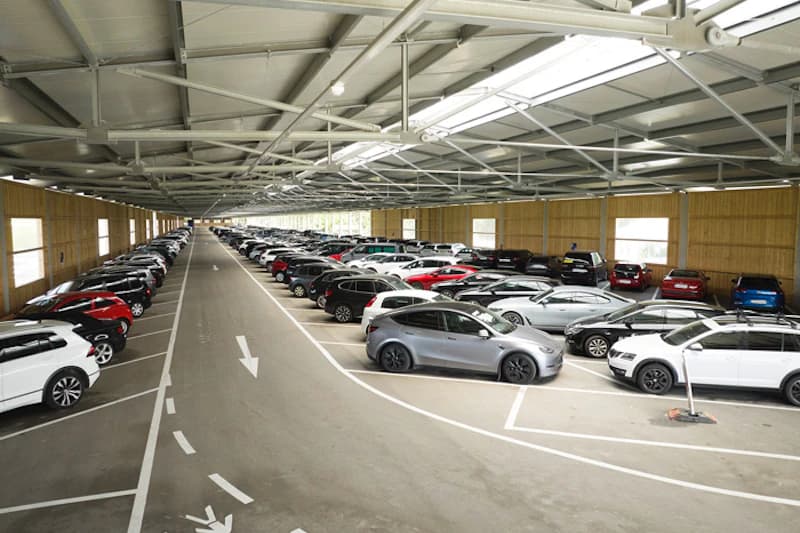 Blick durch eine lange Parkhalle mit vielen geparkten Autos unter einem Stahldach mit Lichtbändern.