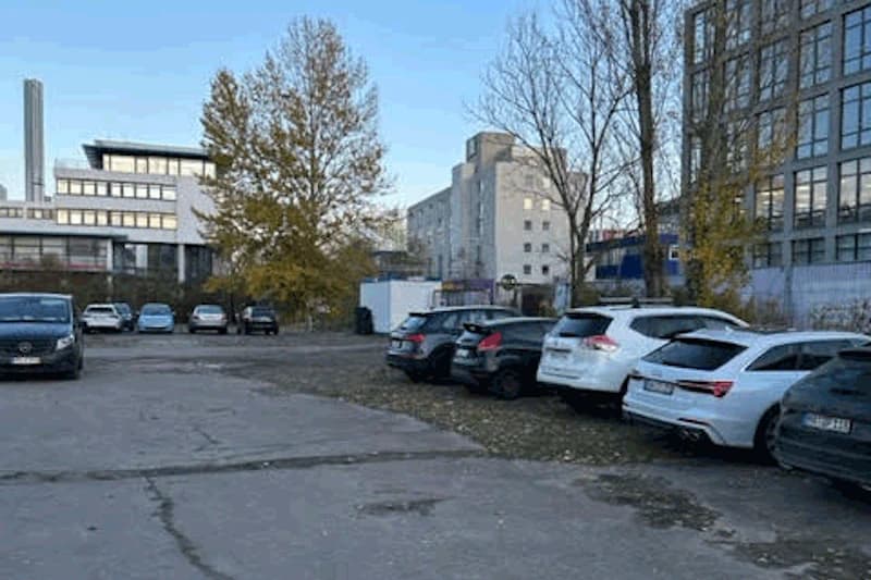 Freier Parkplatz mit in Reihe geparkten Autos, Asphaltfläche, kahle Bäume und mehrstöckige Bürogebäude im Hintergrund.
