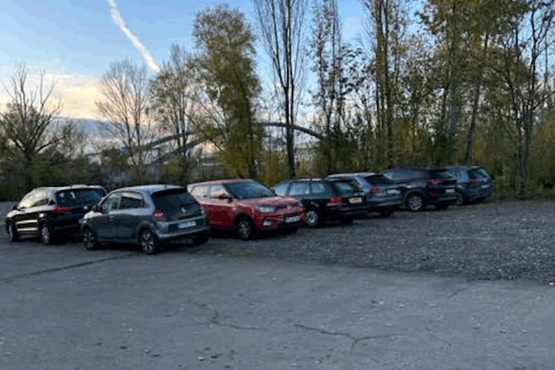 Reihe geparkter Autos auf einem Schotterplatz vor einer Baumreihe und Abendhimmel.