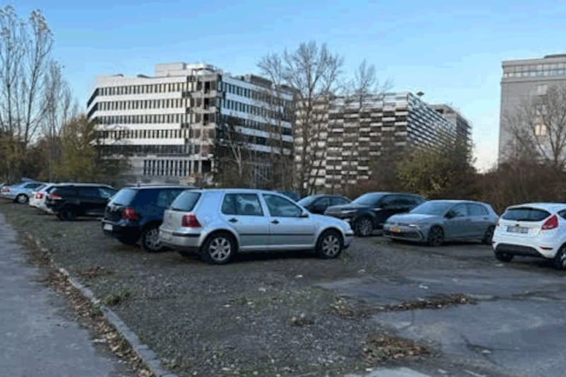 Mehrere geparkte Autos auf einem Schotterparkplatz vor mehrstöckigen Bürogebäuden und kahlen Bäumen.