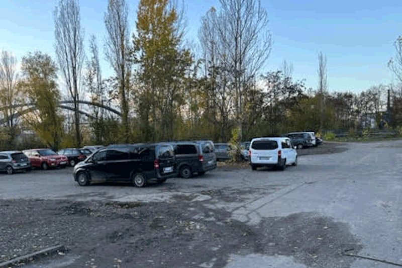 Großer unbefestigter Parkplatz mit mehreren geparkten Vans und Autos, Bäume und eine Brücke im Hintergrund.