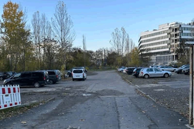 Großes Außenparkplatz mit Autos rechts und links, rissiger Asphalt, Bäume im Hintergrund und mehrstöckiges Gebäude rechts.