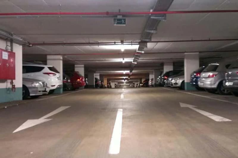 Blick in eine beleuchtete Tiefgarage mit Parkreihen, Pfeilmarkierungen und geparkten Autos.