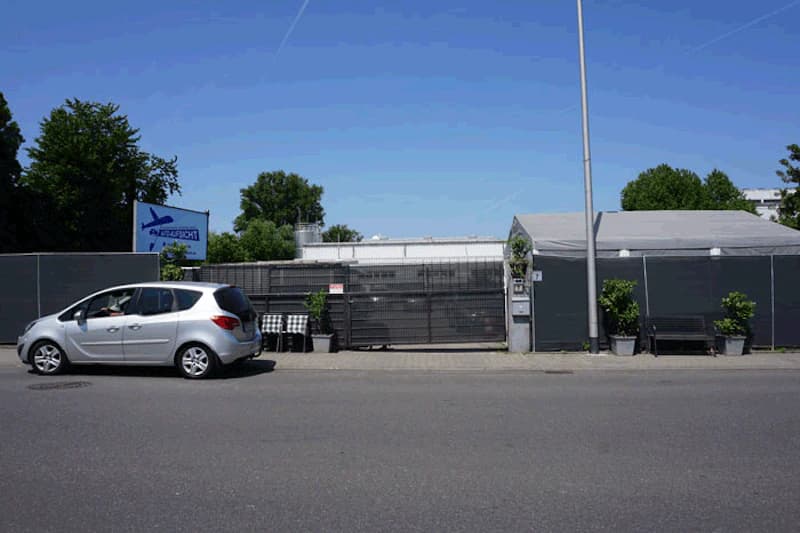 Straße vor eingezäuntem Parkplatz mit geschlossenem Tor, silbernem Auto links und Bänken und Topfpflanzen.