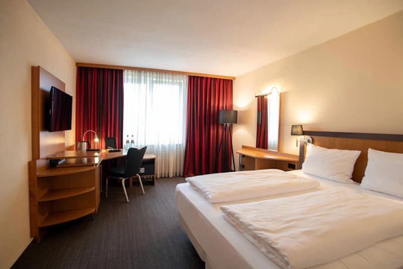 Helles Doppelzimmer mit zwei Einzelbetten, Schreibtisch, Stuhl, Fernseher und roten Vorhängen.
