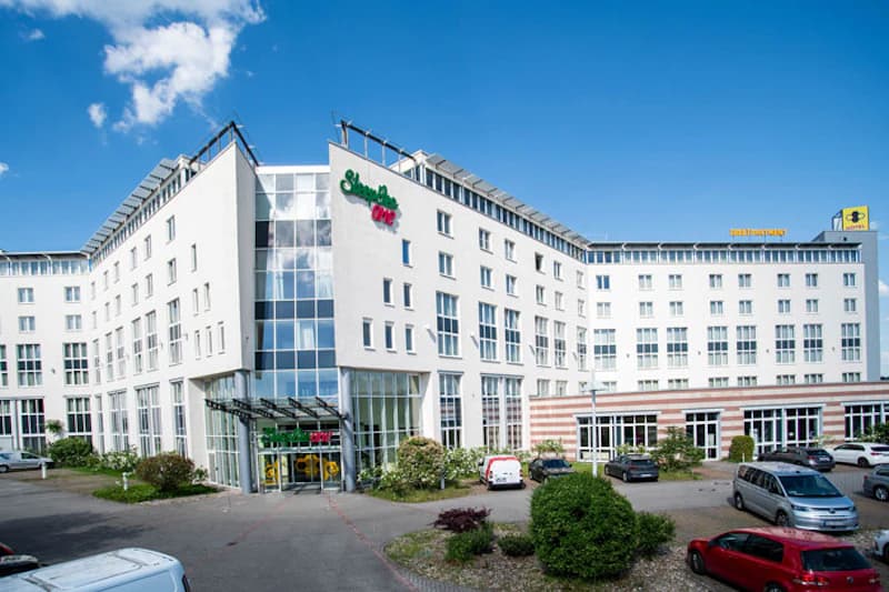 Weißes mehrstöckiges Hotelgebäude mit Glasvorbau, Eingangsbereich, Parkplätzen und Autos unter blauem Himmel.