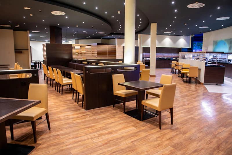 Großer Speisesaal mit Holzboden, dunkelbraunen Tischen, hellen Stühlen und Sitznischen unter Deckenbeleuchtung.