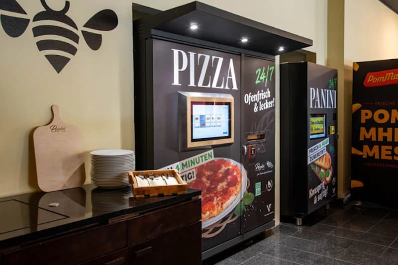 Pizzaautomat und Paniniautomat in Hotelflur neben Stapel Tellern und Besteck.