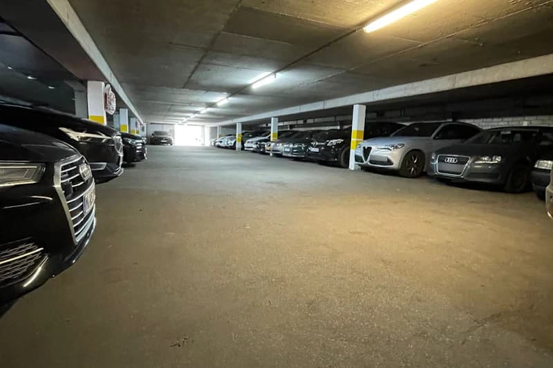 Beleuchtete Tiefgarage mit Reihen geparkter Autos und Stützpfeilern mit gelben Markierungen.