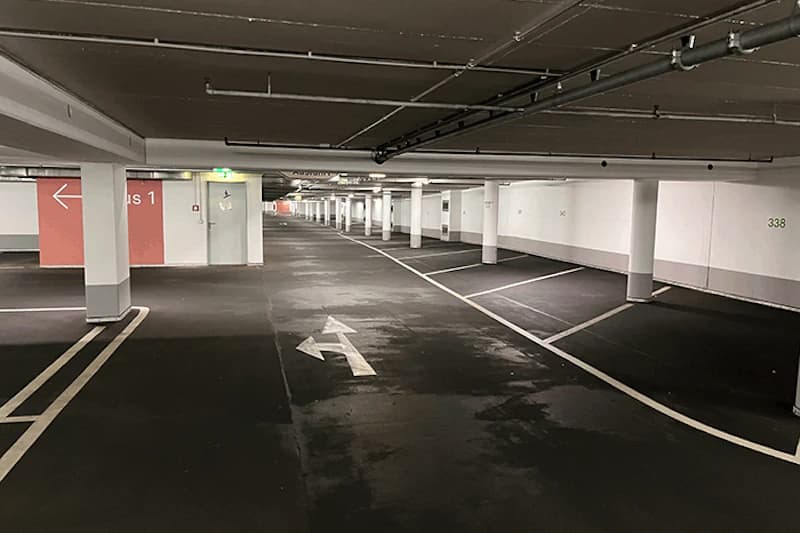 Leere Tiefgarage mit markierten Parkbuchten, Pfeil auf Fahrbahn und beleuchtetem Notausgang.