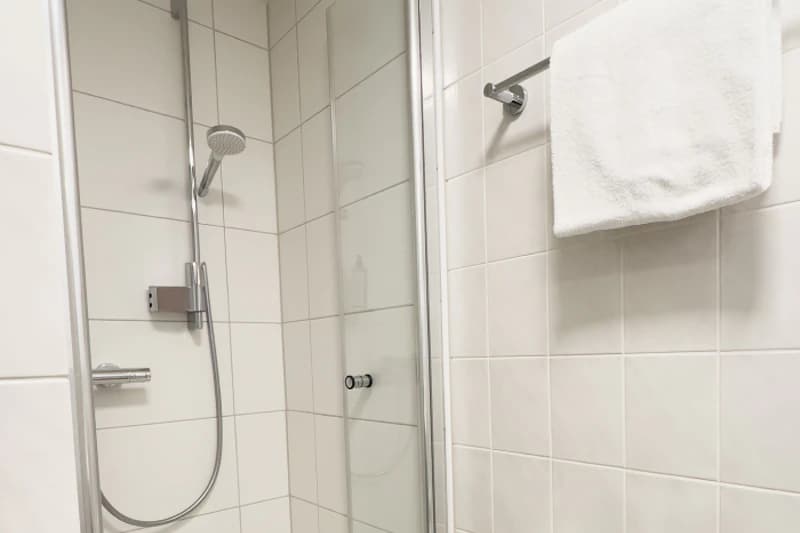 Weiss geflieste Dusche mit Glastür, verstellbarer Handbrause und Handtuch an Wandstange.