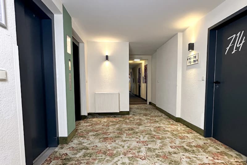 Hotelflur mit gemustertem Teppich, Aufzugstüren links, Zimmertür 714 rechts, Wandlampen und Heizkörper.