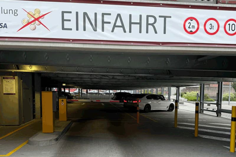 Einfahrt zu einem mehrstöckigen Parkhaus mit Schranke, Stützen und einem weißen Auto im Inneren.