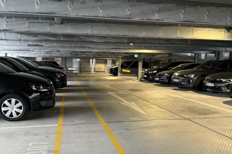 Überdachtes Parkdeck mit geparkten schwarzen Autos, gelben Fahrbahnmarkierungen und Stützsäulen unter niedriger Decke.