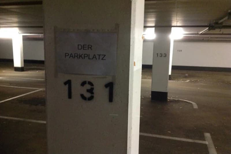 Säule in Tiefgarage mit Schild DER PARKPLATZ und schwarze Zahl 131, leere Parkplätze und weitere Säulen im Hintergrund.